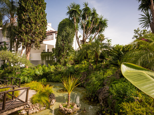 Jardines tropicales en Puente Romano, con exuberante vegetación, un pequeño estanque con patos y una arquitectura tradicional encalada al fondo.