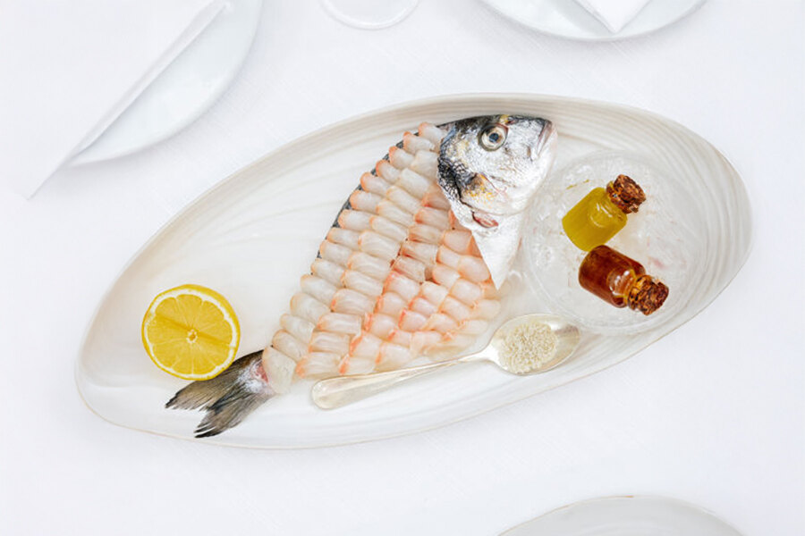 Elegante plato de pescado estilo sashimi servido en Gaia Marbella, con pescado crudo delicadamente cortado sobre el cuerpo de una dorada entera. Se sirve en un plato ovalado blanco con limón, condimentos y dos botellitas de salsa con hielo, todo sobre un mantel blanco impecable.