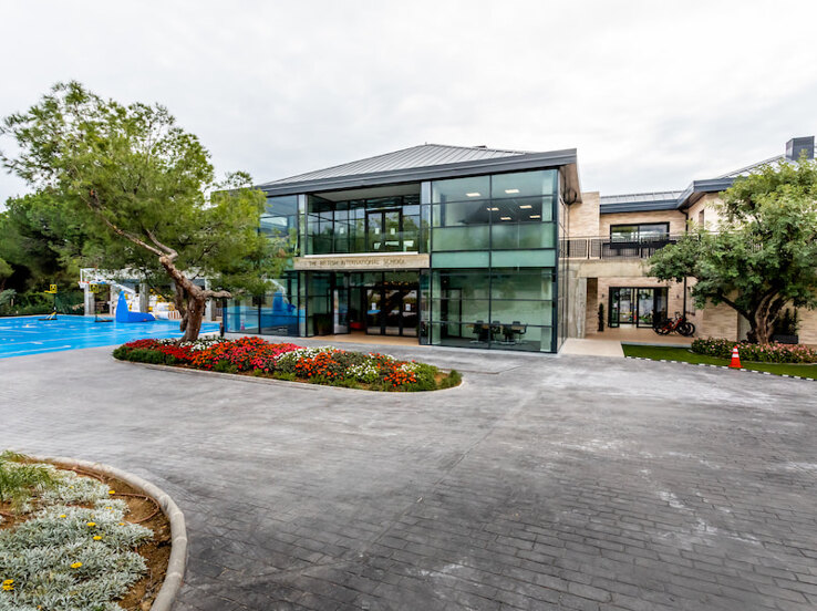 Exterior view of The British International School of Marbella, featuring a modern glass-fronted building surrounded by landscaped gardens, colourful flower beds, and sports facilities. The setting conveys a welcoming and contemporary educational environment.