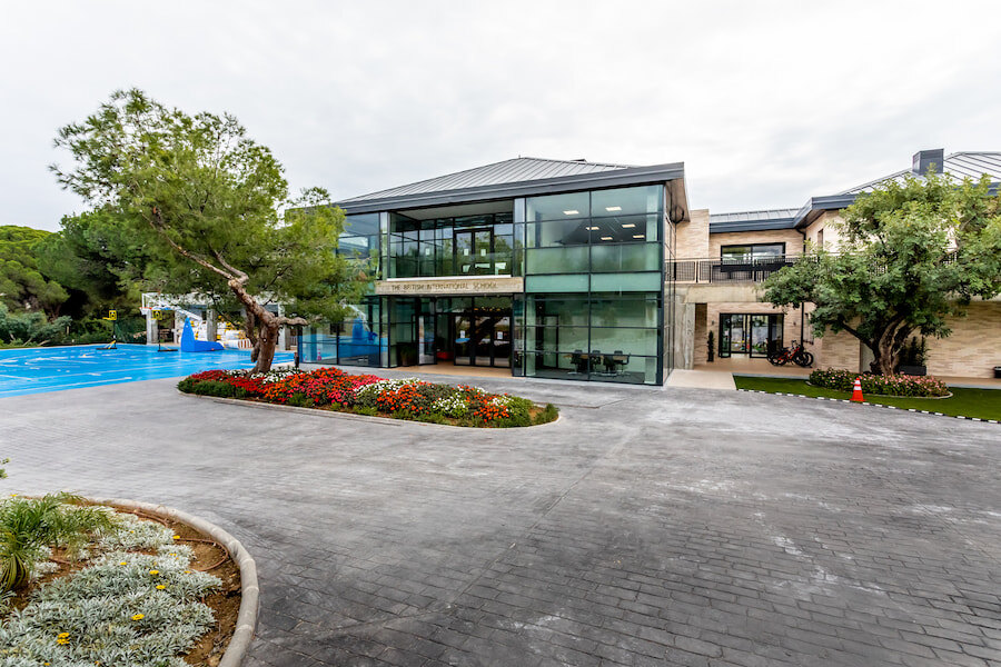 Exterior view of The British International School of Marbella, featuring a modern glass-fronted building surrounded by landscaped gardens, colourful flower beds, and sports facilities. The setting conveys a welcoming and contemporary educational environment.