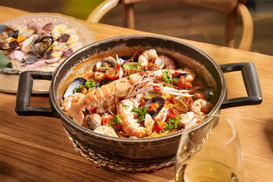 Primer plano de un plato de mariscos servido en una sartén rústica en El Chiringuito Marbella, con gambas, almejas, mejillones y langostinos en una rica salsa de tomate con hierbas. Al fondo, un plato de mariscos variados y una copa de vino blanco completan la experiencia gastronómica mediterránea.