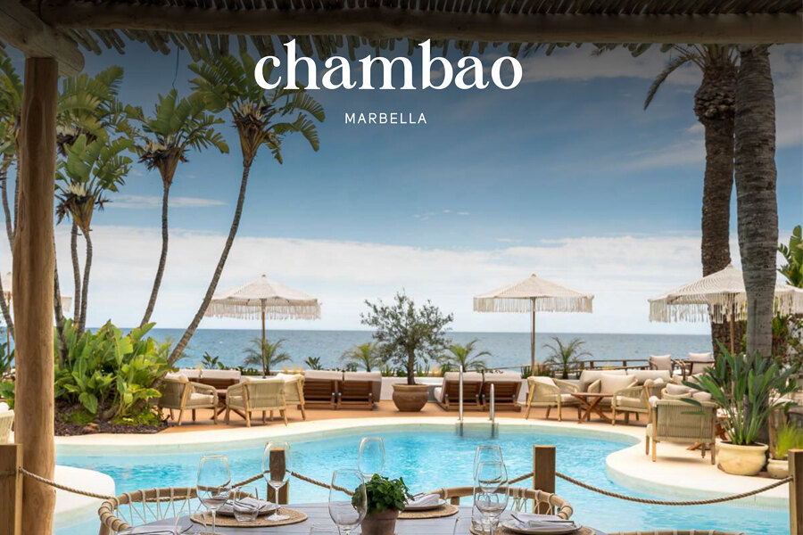 Vista panorámica desde Chambao Marbella en Puente Romano, con un elegante comedor frente al mar con piscina circular, palmeras tropicales y sombrillas de paja. Las mesas están servidas con cristalería y rodeadas de muebles de madera natural, con vistas al mar Mediterráneo bajo un cielo azul.