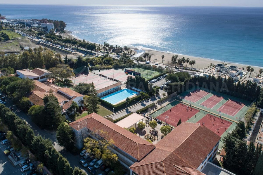 Schools in Marbella. Aerial view of Colegio San José in Estepona, located along the Mediterranean coast, featuring red-roofed buildings, multiple sports courts, a swimming pool, and direct access to the beach. The school is surrounded by greenery and offers stunning sea views, highlighting its privileged seaside setting.