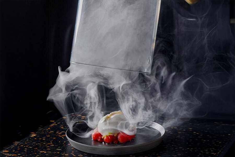 Una presentación culinaria espectacular en Leña Marbella, con un plato ahumado desvelado bajo una cúpula de cristal. Espirales de humo se elevan para revelar una creación cuidadosamente presentada con texturas cremosas y vibrantes frutos rojos, sobre un fondo oscuro y elegante.