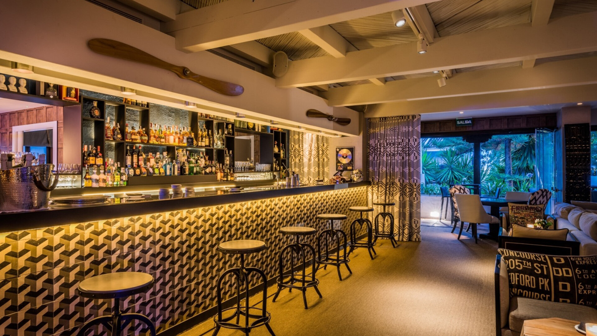 Owner´s club. Stylish cocktail bar at Puente Romano, featuring geometric-patterned tiles, a sleek illuminated counter, and an extensive selection of premium spirits. The warm lighting and eclectic decor create a cosy, sophisticated atmosphere with views of lush tropical gardens through large windows.