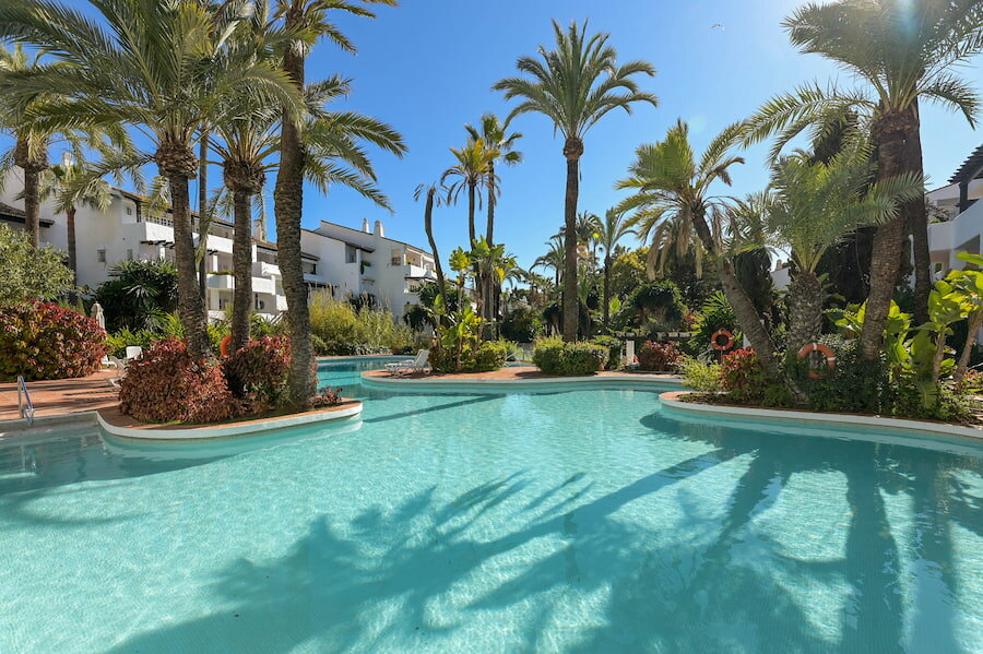 Swimming pool surrounded by tropical gardens and white Mediterranean-style buildings in Puente Romano Phase II residential complex in Marbella.