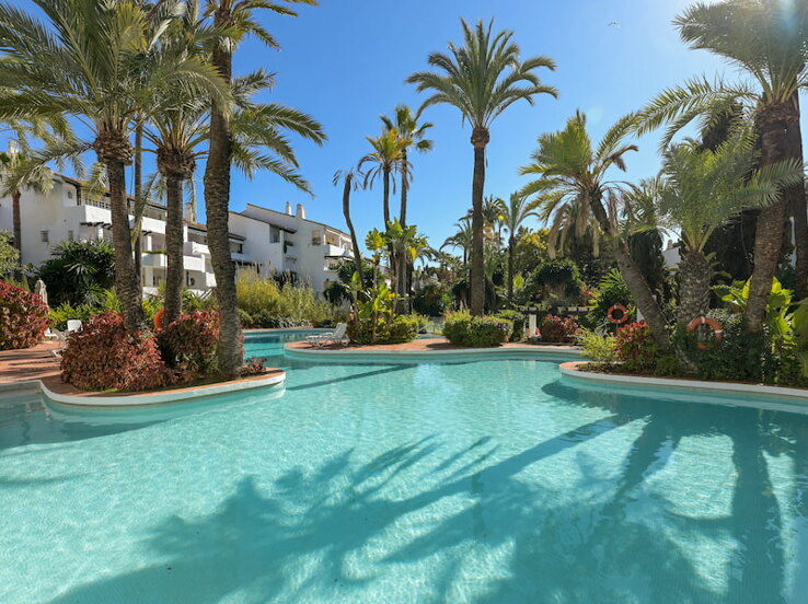 Piscina rodeada de jardines tropicales y edificios blancos de estilo mediterráneo en el complejo residencial Puente Romano Fase II en Marbella.