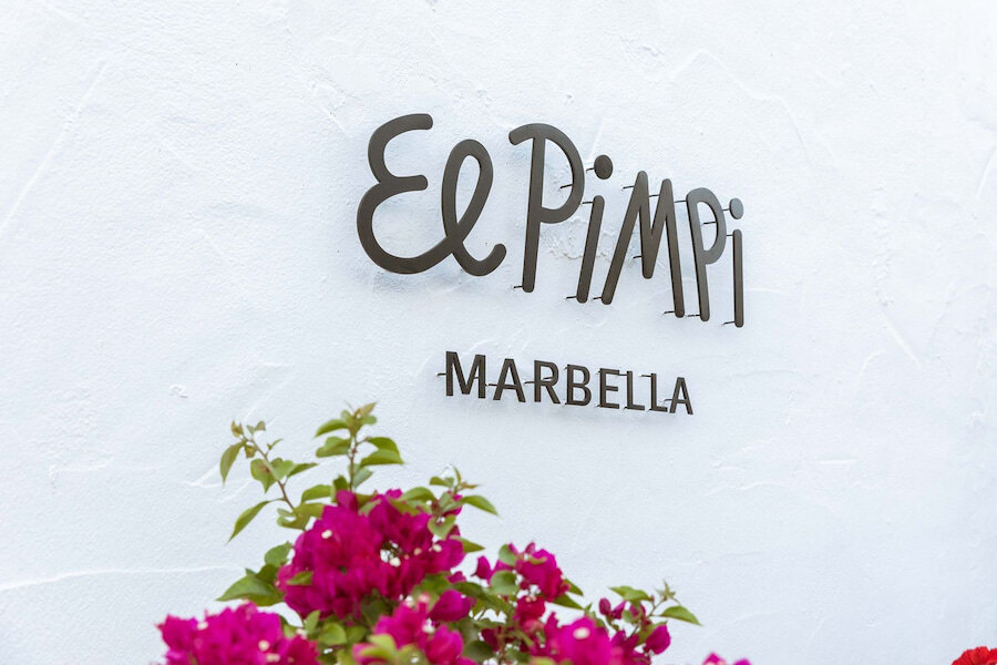 Interior de El Pimpi Marbella, ubicado en Puente Romano, con un comedor luminoso y elegante con sillas tapizadas a rayas, mesas de mármol y un encantador mural con pavos reales azules y motivos florales. La vegetación vertical y la suave luz natural crean un ambiente andaluz fresco y acogedor.