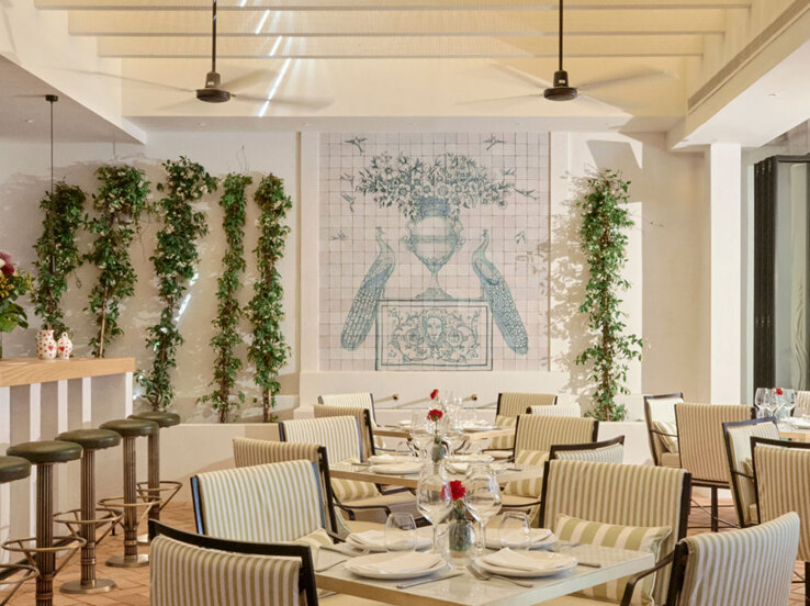 Interior de El Pimpi Marbella, ubicado en Puente Romano, con un comedor luminoso y elegante con sillas tapizadas a rayas, mesas de mármol y un encantador mural con pavos reales azules y motivos florales. La vegetación vertical y la suave luz natural crean un ambiente andaluz fresco y acogedor.