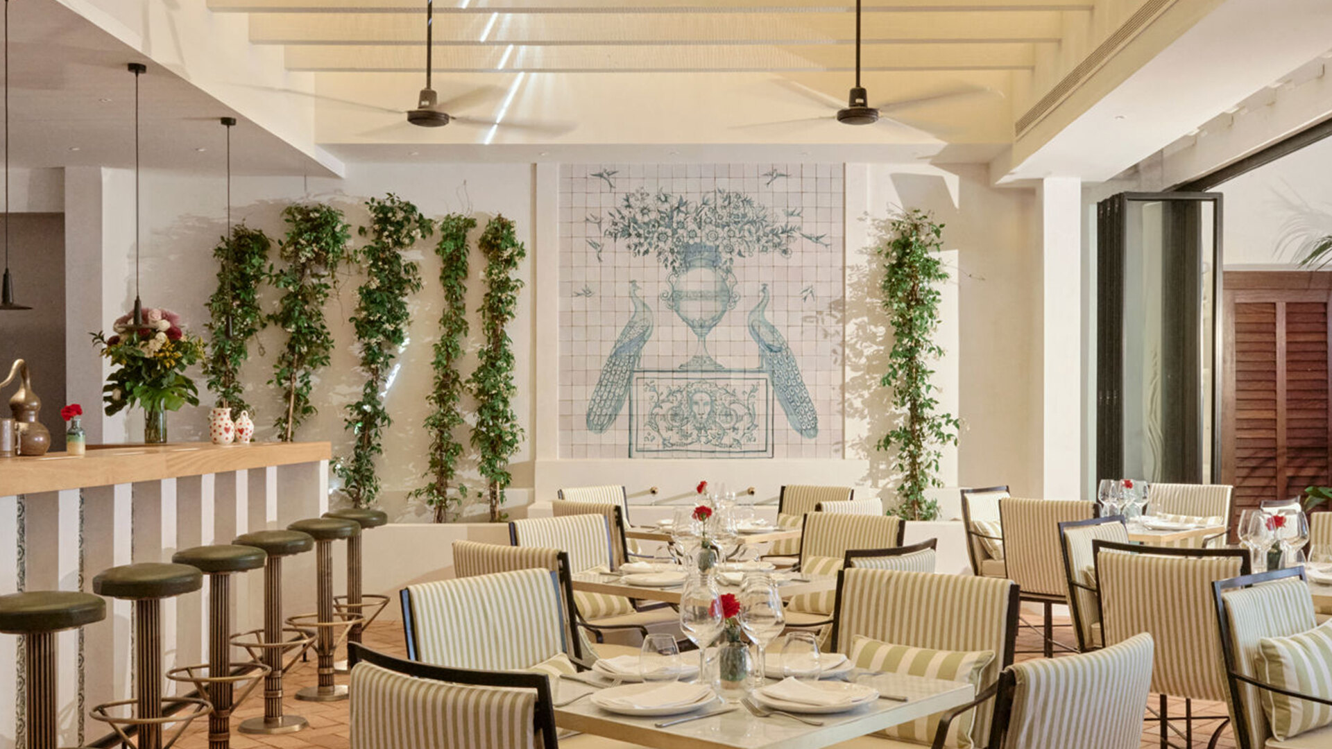Interior de El Pimpi Marbella, ubicado en Puente Romano, con un comedor luminoso y elegante con sillas tapizadas a rayas, mesas de mármol y un encantador mural con pavos reales azules y motivos florales. La vegetación vertical y la suave luz natural crean un ambiente andaluz fresco y acogedor.