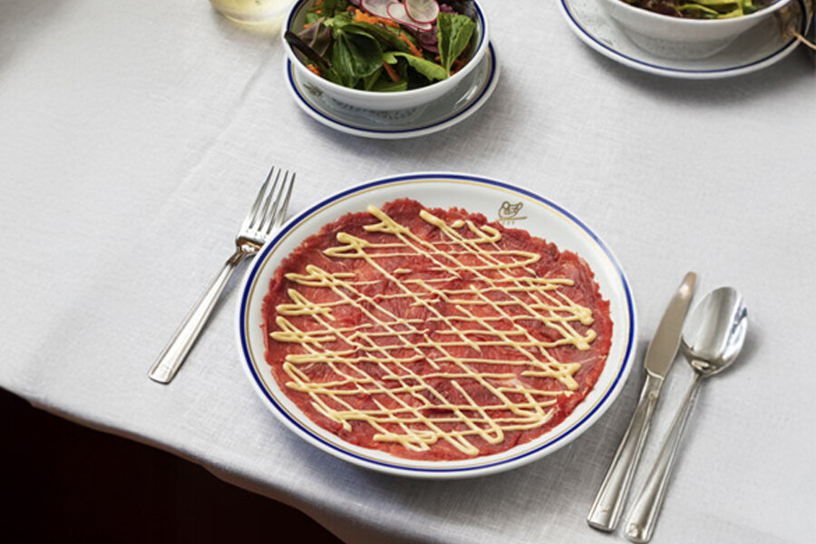 Carpaccio de ternera elegantemente presentado con un toque de cremoso aderezo en Cipriani Marbella, ubicado en el exclusivo resort Puente Romano. El plato se sirve sobre un mantel blanco, acompañado de una ensalada fresca y una copa de vino blanco, creando un ambiente refinado y sofisticado.