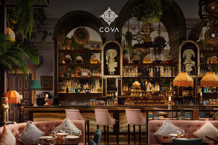Interior de COYA Marbella, ubicado en Puente Romano, que presenta un elegante comedor con cómodos asientos, decoración artística y un vibrante bar enmarcado por espejos arqueados y vegetación tropical. La cálida iluminación, el mobiliario ecléctico y las ricas texturas crean un ambiente sofisticado y acogedor.