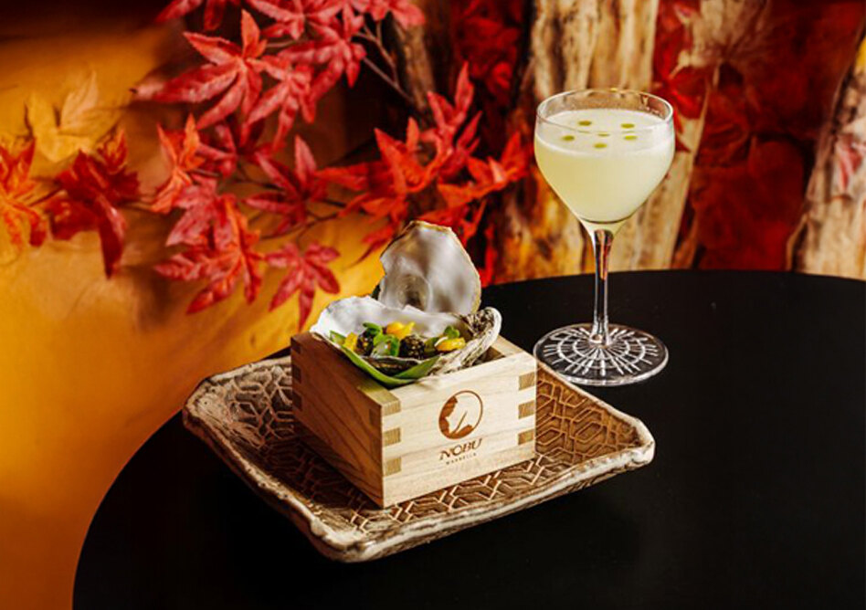 Plato gourmet en Nobu Marbella, presentado en bandeja con una caja de madera grabada con el logotipo del restaurante, que contiene una ostra con ingredientes frescos. Junto a ella, una elegante copa de cóctel se alza sobre un fondo otoñal de hojas rojas y tonos cálidos.