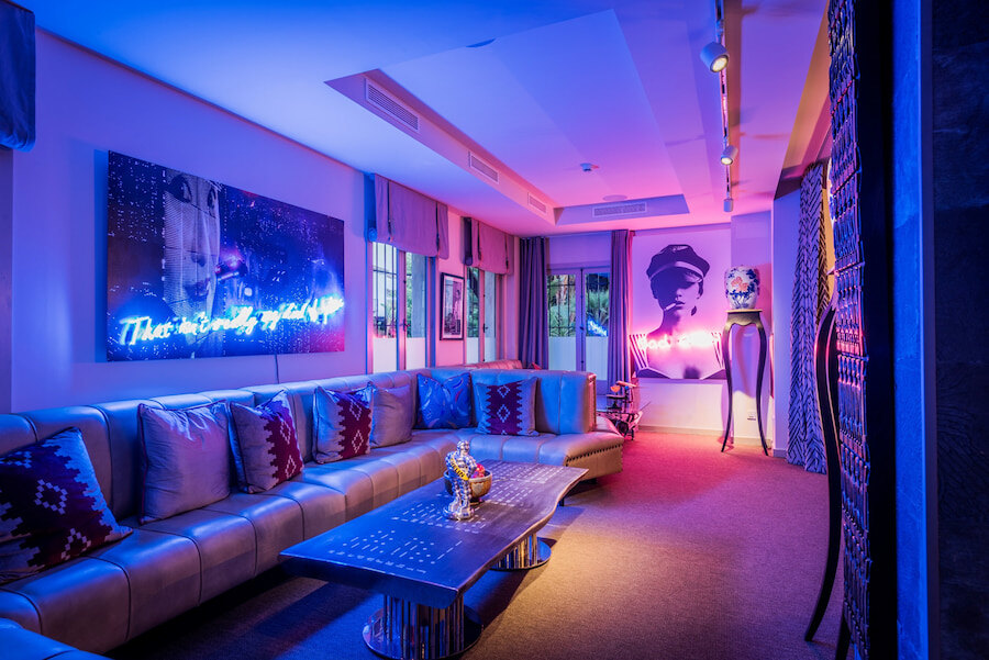 Interior of the Owner’s Club at Puente Romano, featuring a sophisticated contemporary lounge with neon ambient lighting, a leather L-shaped sofa, modern artwork, and exclusive décor in a vibrant, elegant setting.
