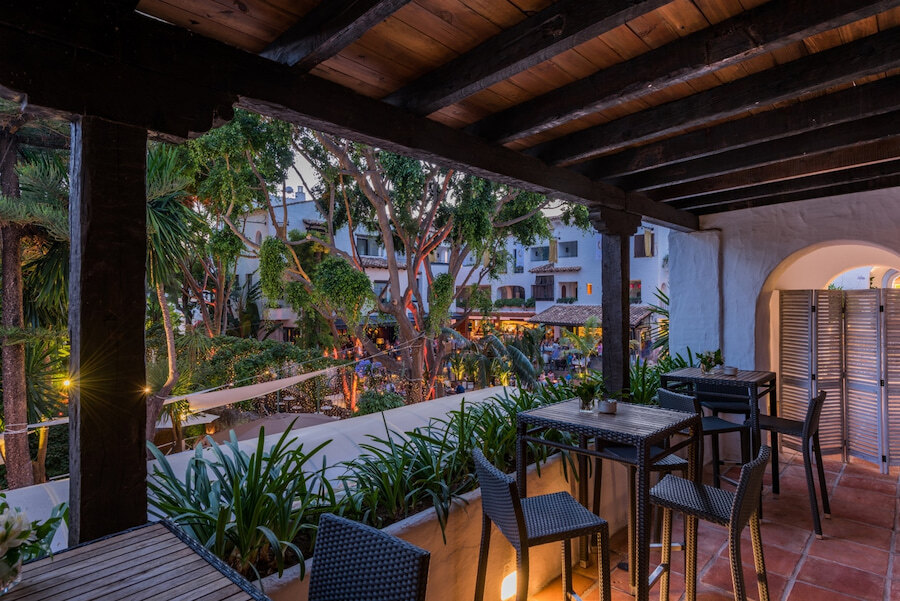 Terrace of the Owner’s Club at Puente Romano Marbella, overlooking the illuminated gardens and the lively central plaza of the resort. A cosy space with high tables, lush greenery, and traditional Andalusian architecture.