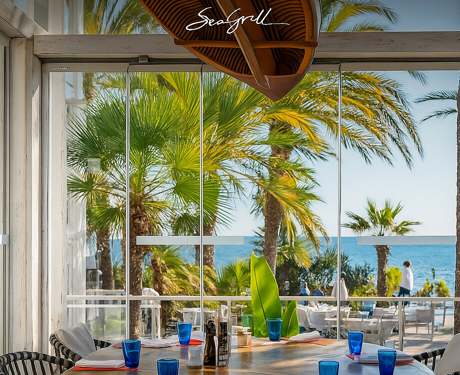 Restaurante Sea Grill: Interior del restaurante Sea Grill con una mesa redonda elegantemente dispuesta, copas azules y vistas al mar Mediterráneo a través de amplios ventanales enmarcados por palmeras. Al fondo, se aprecia la terraza con sombrillas blancas y el mar azul bajo un cielo despejado.