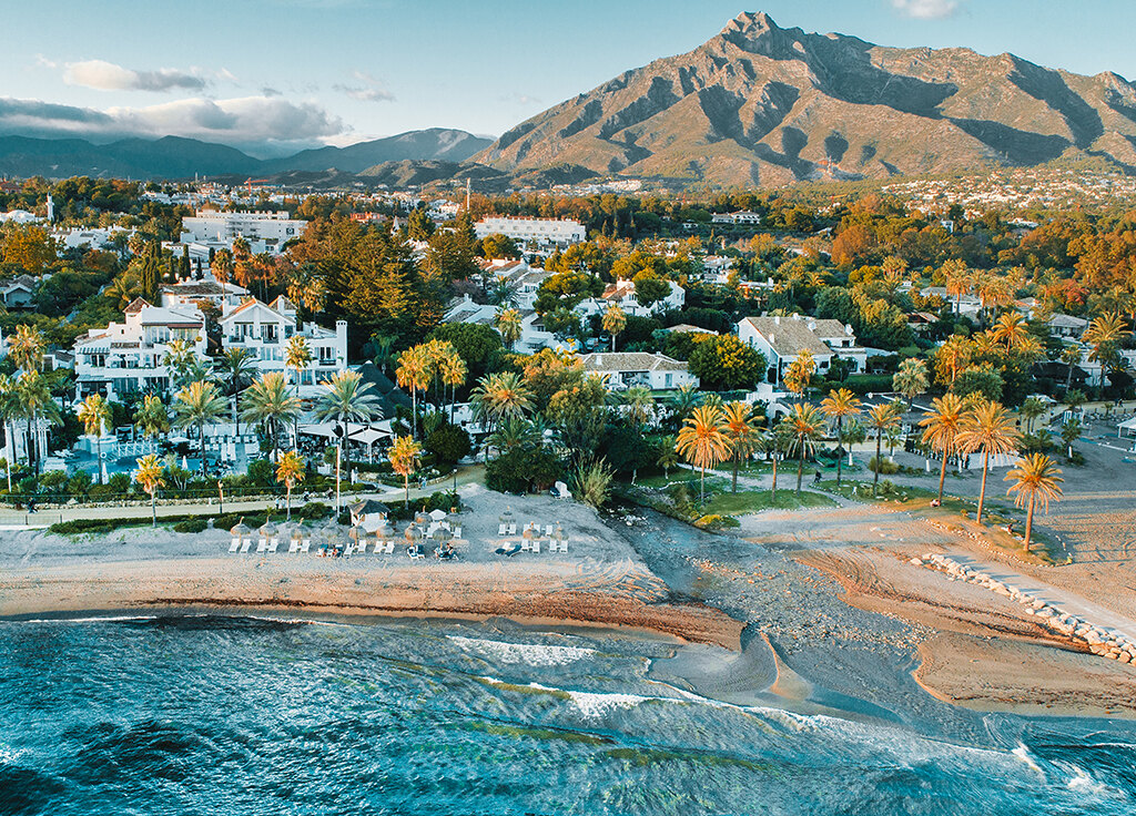 Vista aérea de la zona de Puente Romano en Marbella, con villas frente a la playa, exuberantes palmeras y la icónica montaña La Concha al fondo.