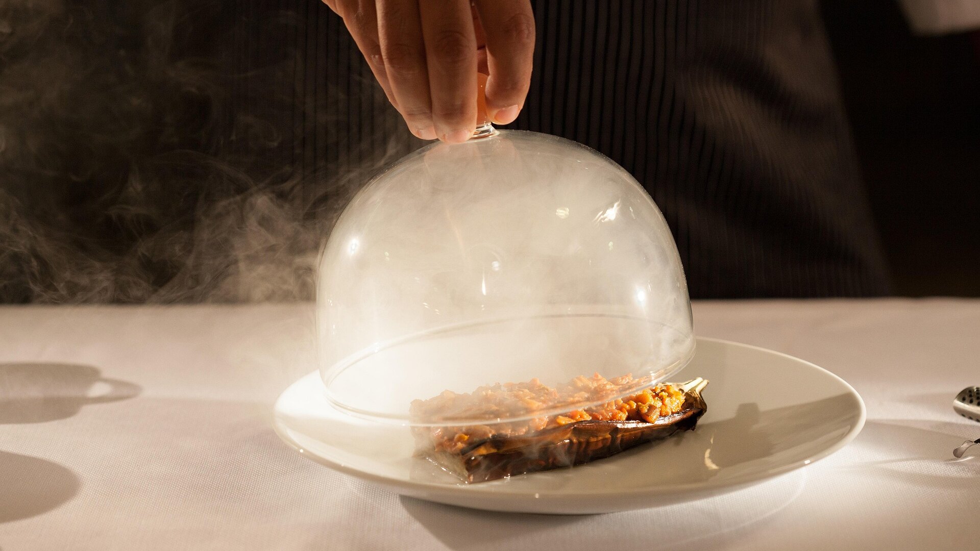 Plato gourmet servido bajo una campana de cristal con humo, siendo descubierto por la mano del camarero.